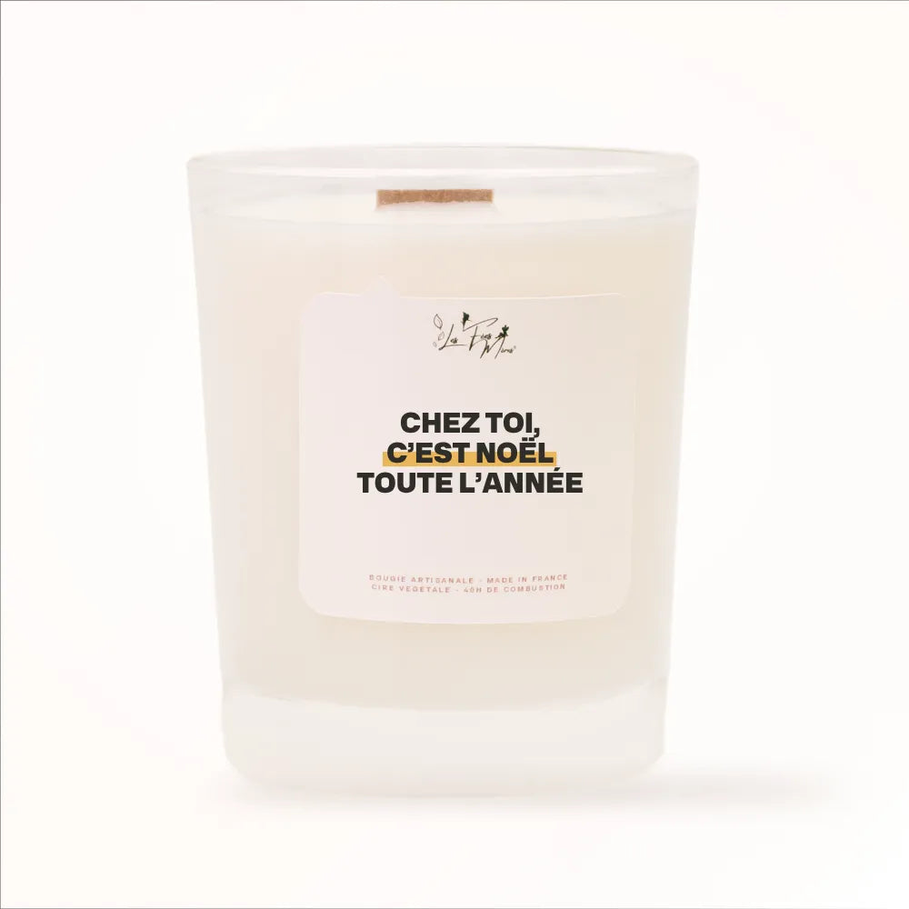 Bougie parfumée de Noël Les Fées Mères avec l'étiquette « Chez toi, c'est Noël toute l'année », pot blanc et mèche en bois de hêtre