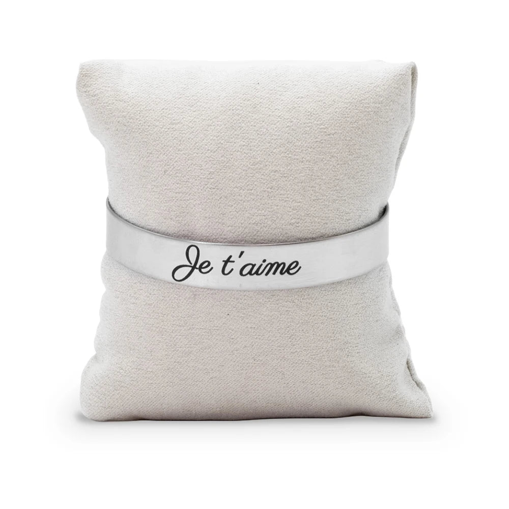 Bracelet jonc gravé Je t'aime en écriture manuscrite noire sur acier inoxydable, présenté sur un coussin en tissu beige, Les Fées Mères